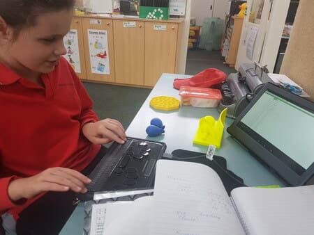 A primary-school age student sits at a desk, using a braille writer device and a screen that translates it for teachers. She is surrounded by colourful learning aids and posters in classroom.