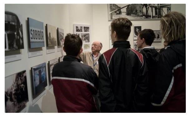 An older man speaks to a group of male secondary school students, showing them documentary photos on the wall at the Jewish Holocaust Centre