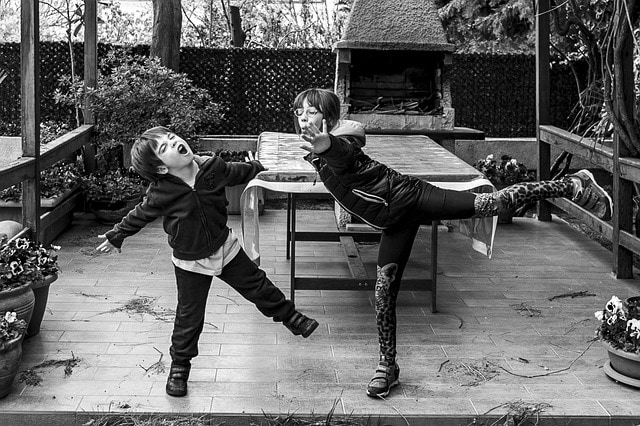 Two children act out a dramatic game outdoors, one balancing on one leg like a plane with arms out, the other reacting with eyes closed and face screwed up.