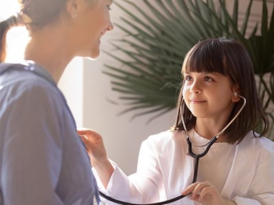 A smiling child dresses up as a doctor using a stethoscope and a white coat.