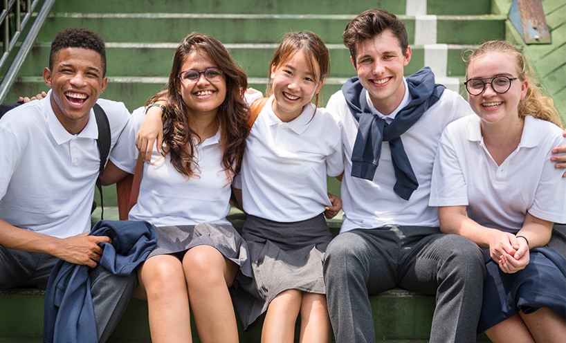 Five high-school students in white and grey uniforms sitting on the stairs and smiling towards the camera.