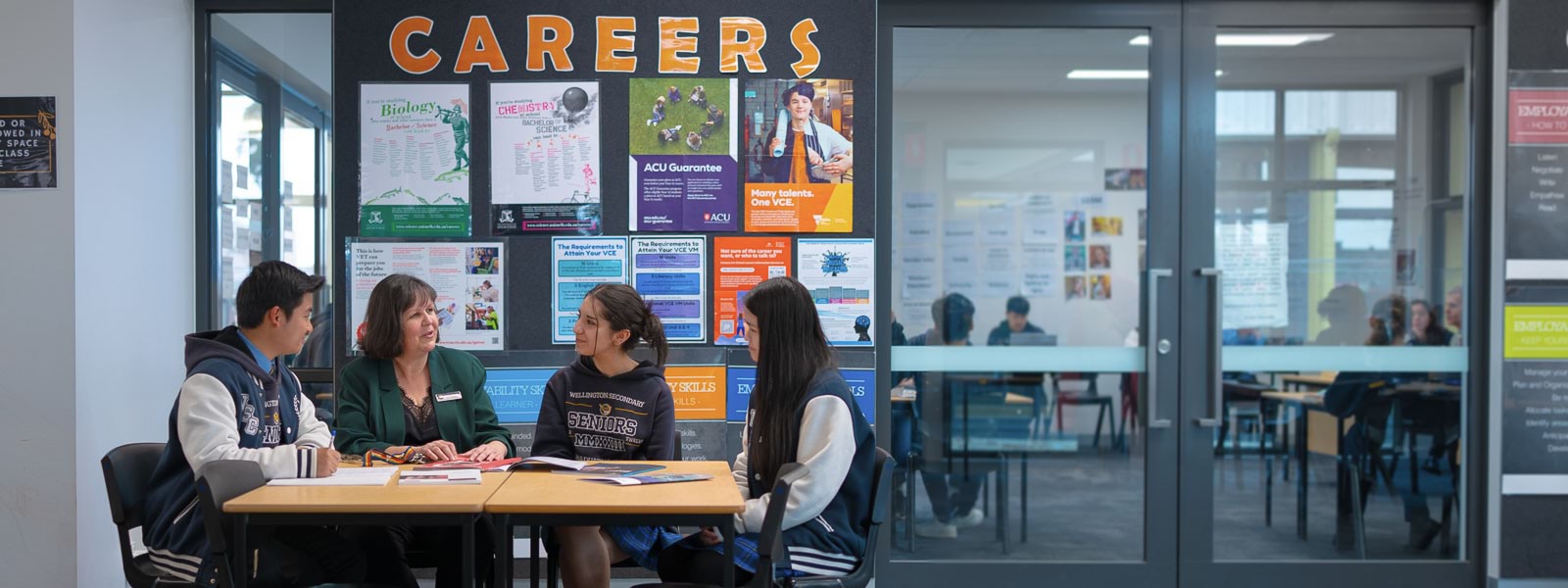 A woman and 3 students sitting at a table. Behind them is a pin board with career information sheets.